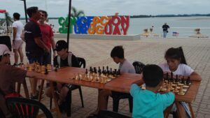 Imagen de jóvenes jugando al ajedrés en la playa. De fondo se observa arena; playa y un cartel colorido de Concepción del Uruguay